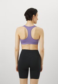 Ljubičasti atletski top s dizajnom racerback, uparen s crnim šortsevima visokog struka koji imaju mali Nike logo na stražnjoj strani.