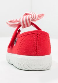 Sneaker di tela rossa vista posteriore con suola in gomma bianca e fiocco in tessuto rosa e bianco a righe annodato alla linguetta del tallone.