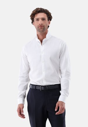 Mann mit lockigem Haar, trägt ein weißes Hemd mit Stehkragen, das in schwarze Hosen mit schwarzem Gürtel gesteckt ist, steht vor einem schlichten Hintergrund.