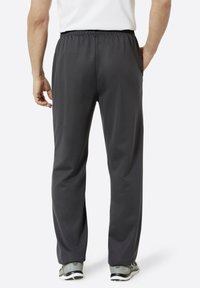 Mann trägt dunkelgraue elastische Freizeit-Hose und graue Sneakers, von hinten betrachtet vor einem schlichten weißen Hintergrund.