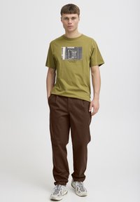 T-shirt in cotone verde oliva con stampa fotografica sul davanti, abbinata a pantaloni marroni larghi e sneakers grigie.