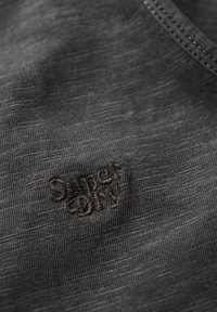 Ciemnoszara tkanina z subtelną teksturą, z haftowanym logo "Super Dry" w ciemniejszym odcieniu, umieszczonym w dolnym lewym rogu.