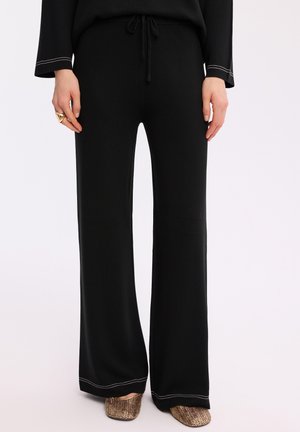 Kobieta w czarnych szerokich spodniach z białym przeszyciem i złotych, teksturowanych płaskich butach, stoi z rękami swobodnie opuszczonymi wzdłuż ciała.