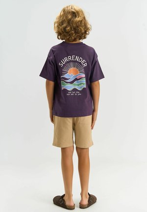 Barn med krøllet blondt hår iført en lilla t-shirt med en solnedgangsgrafik og ordet "SURRENDER", beige shorts og brune sandaler, stående.