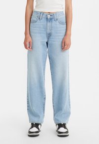 Kvinna klädd i ljusblå högmidjade, löst sittande jeans, vit kort t-shirt och svart-vita sneakers, stående mot vit bakgrund.