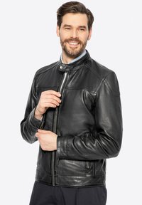 Uomo sorridente che chiude la giacca di pelle nera sopra una camicia blu contro uno sfondo bianco uniforme.