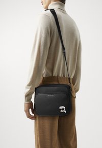 Borsa a tracolla nera con esterno testurizzato, tracolla regolabile e grafica di cartone sul davanti. Chiusura con zip e dettagli di branding.