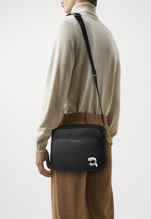 Black crossbody bag with textured exterior, adjustable strap, and a cartoon graphic on the front. Zippered closure and branded detailing.