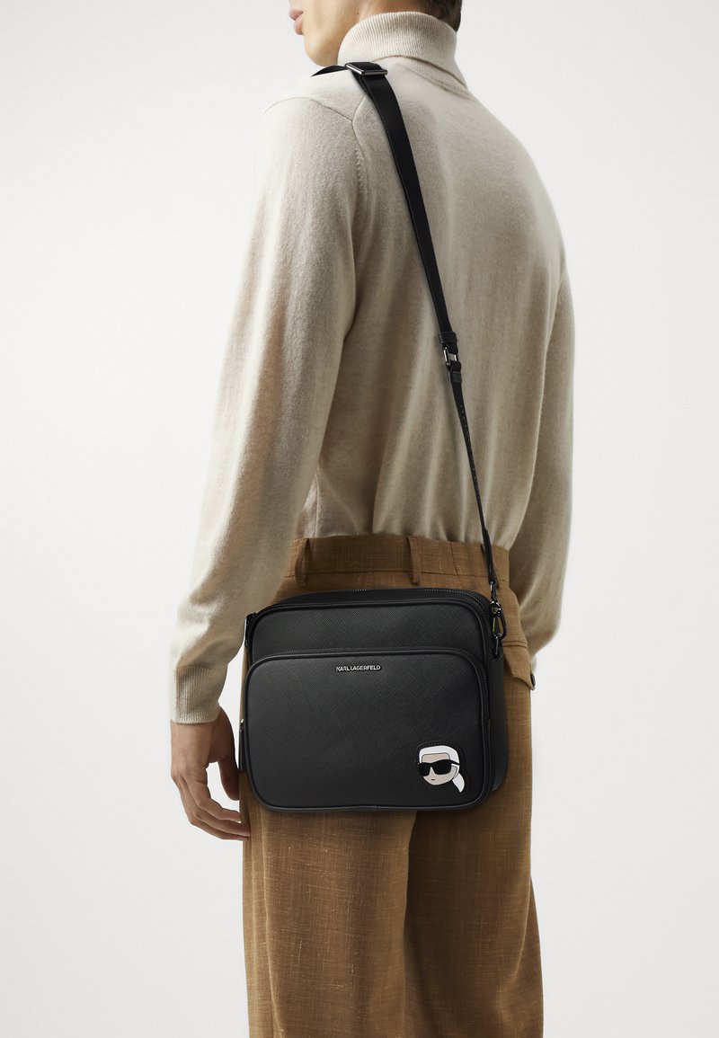 Borsa a tracolla nera con esterno testurizzato, tracolla regolabile e grafica di cartone sul davanti. Chiusura con zip e dettagli di branding.