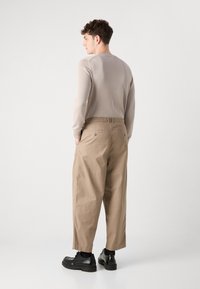 Beige long-sleeve shirt and loose-fitting khaki trousers. Black shoes with a chunky sole. Back view showing pockets and relaxed silhouette.