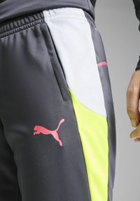 Sive trenirke s bijelim mrežastim panelom i neon žutim detaljima. Imaju ružičasti Puma logo na lijevom bedru i detalje s patentnim zatvaračem.