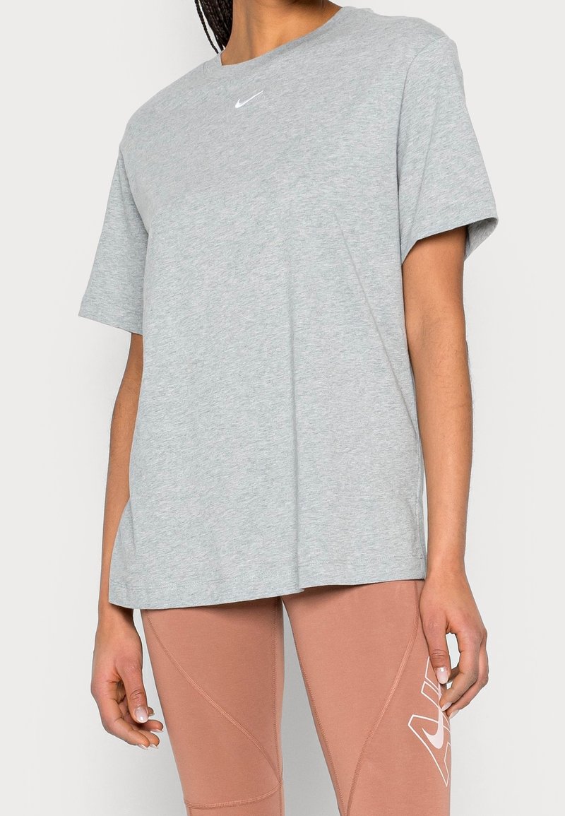 Person som bär en ljusgrå Nike-t-shirt och rosa leggings med vit logotyp, stående mot en plain ljus bakgrund.