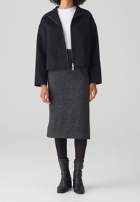 Femme portant une veste zippée noire courte, une chemise blanche, une jupe midi grise, des collants noirs et des bottines à talons noires, debout devant un fond blanc.