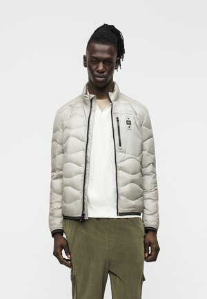 Bărbat cu piele închisă la culoare și dreadlocks, purtând o geacă matlasată gri deschis peste un tricou alb și pantaloni din catifea cu o nuanță de verde măsliniu, stând în fața unui fundal simplu.