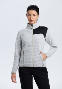 Chaqueta gris con cremallera y un patrón repetido, con un acento negro en el hombro, bolsillos laterales y un cuello alto. Textura suave y diseño ajustado.