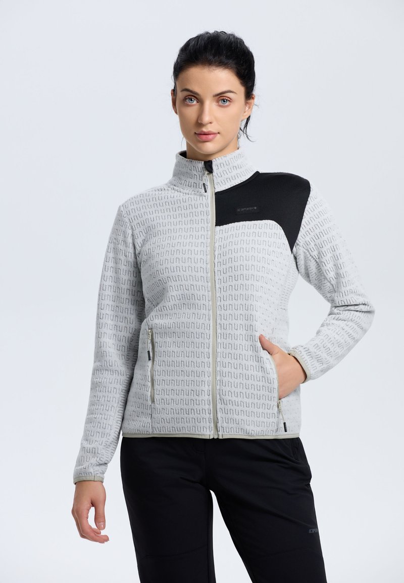 Chaqueta gris con cremallera y un patrón repetido, con un acento negro en el hombro, bolsillos laterales y un cuello alto. Textura suave y diseño ajustado.
