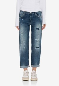 Jeans azules desgastados y recogidos, combinados con zapatillas blancas y una camiseta blanca de manga larga, mostrados de la cintura a los pies.