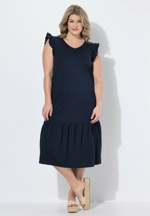 Kvinde står mod hvid væg iført en mørkeblå kjole uden ærmer med flæser på skuldrene og beige plateau-sandaler.