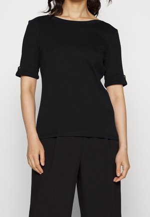 Kobieta ubrana w czarną bluzkę z krótkim rękawem z podwiniętymi mankietami i guzikiem, w połączeniu z luźnymi czarnymi spodniami na neutralnym tle.