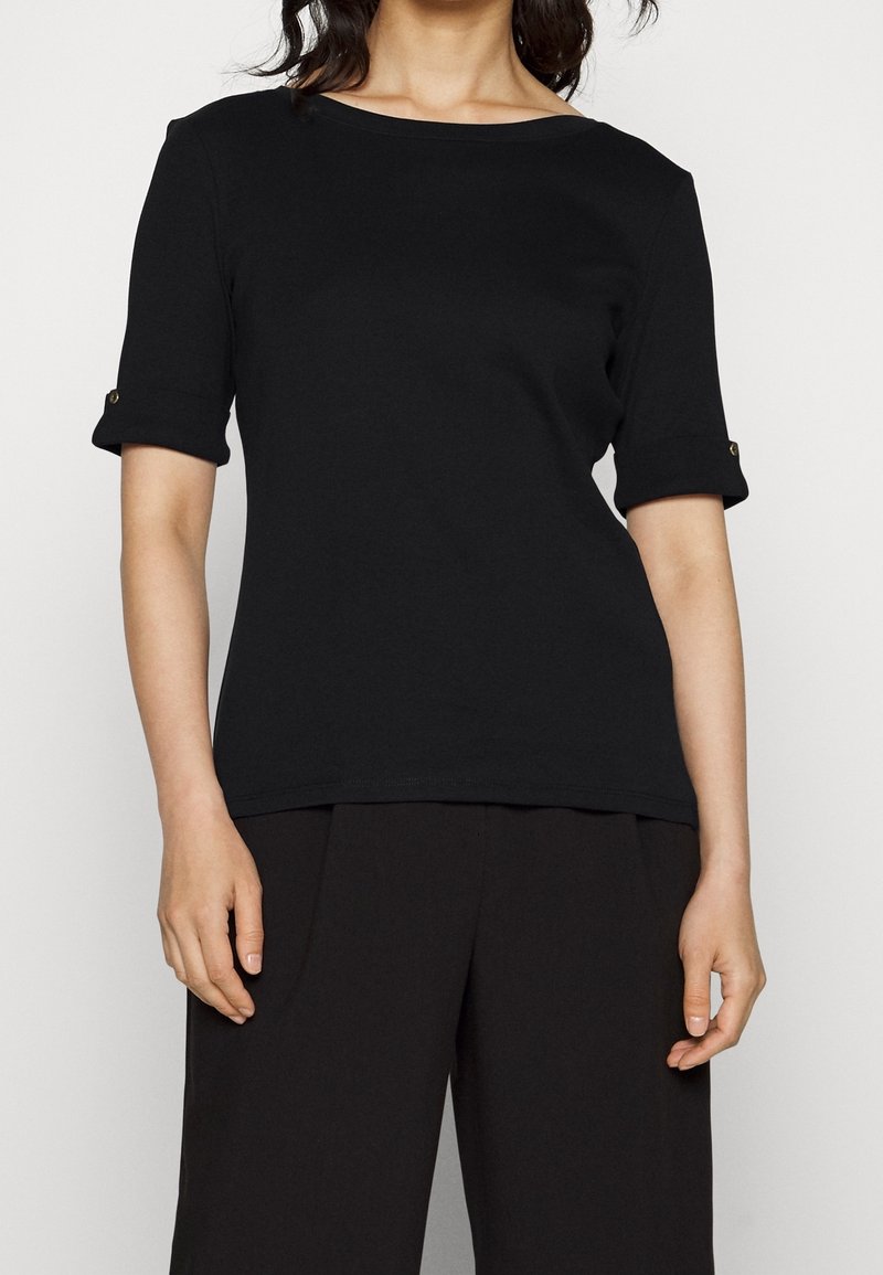 Femme portant un haut noir à manches courtes avec les poignets retroussés et un détail de bouton, associée à un pantalon noir ample sur un fond neutre.