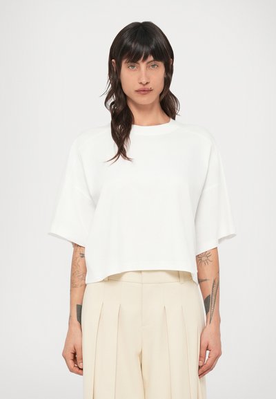 T-shirt blanc court à coupe décontractée, manches courtes et texture lisse, assorti à un pantalon beige plissé.