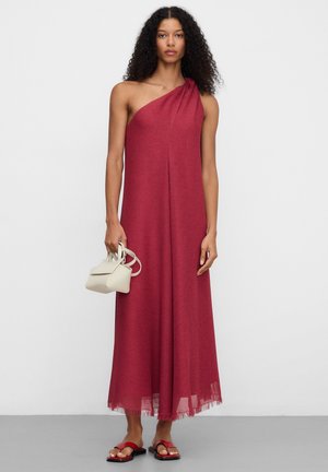 Donna con capelli ricci che indossa un abito rosso lungo, asimmetrico e senza maniche, tiene una piccola borsa beige e indossa sandali rossi.