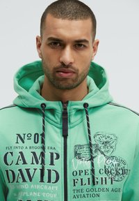 Sudadera con cremallera en verde menta hecha de tela suave, con cordones oscuros, texto impreso y gráficos temáticos de aviación vintage.