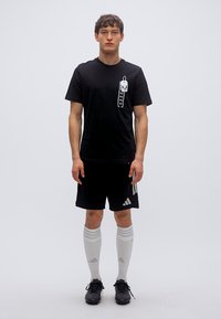 Μαύρο κοντομάνικο T-shirt με γραφικό στο μπροστινό μέρος, συνδυασμένο με μαύρα σορτς με λευκές ρίγες. Λευκές κάλτσες πάνω από το γόνατο και μαύρα παπούτσια.