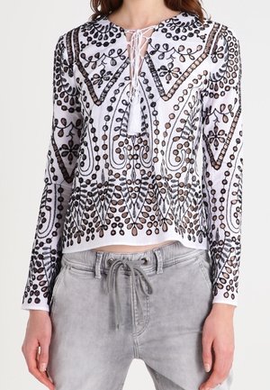 Crop top blanc brodé avec des motifs intricats noirs et bruns, manches longues et un col à nouer avec un détail en tassel.