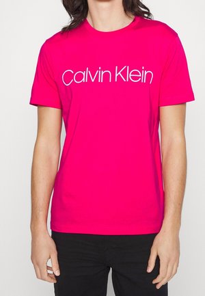 Personne portant un t-shirt à manches courtes rose vif avec le logo blanc "Calvin Klein" sur la poitrine, accompagné d'un pantalon noir.