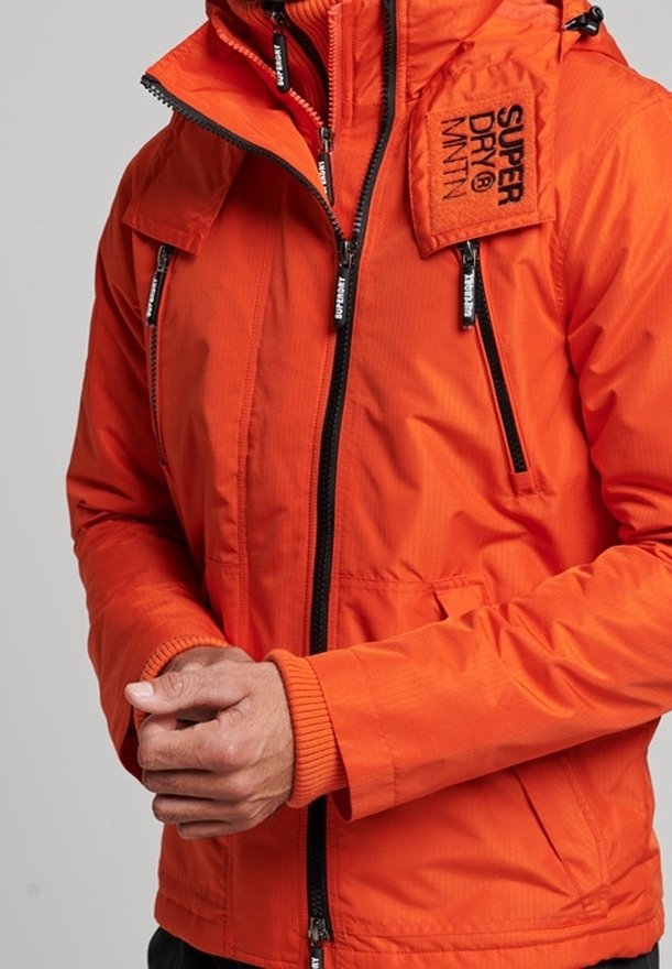 Veste orange vif avec des fermetures éclair noires, marquée "Super Dry MNTN" sur la poitrine, portée par une personne aux mains croisées.
