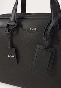 Sort struktureret taske med prægning. Har læderhåndtag og to tags med "BOSS" logo. Slankt, minimalistisk design.