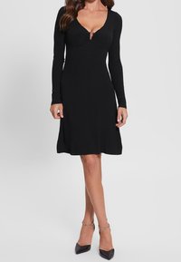 Robe noire côtelée à manches longues, décolleté en V profond, et corsage ajusté s'évasant légèrement à l'ourlet. Portée avec des escarpins noirs à bout pointu.