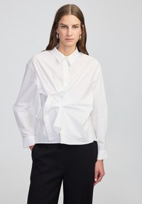 White button-up shirt with long sleeves, featuring a twisted fabric detail at the waist. Smooth cotton texture, classic collar, tailored fit.