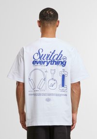 Hvid bomulds T-shirt med blå grafisk tekst og illustrationer på bagsiden, herunder hovedtelefoner og en plan. Korte ærmer, afslappet pasform.