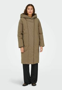Padded long coat w kolorze oliwkowym z kapturem, o minimalistycznym designie i pionowych szwach. Nosi się go na czarnych szerokich spodniach.