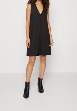 Woman wearing sleeveless black V-neck A-line dress paired with black ankle boots, standing against a plain white background.