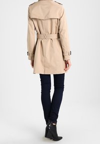 Beige trench coat dengan pinggang yang terikat, epaulet bahu, dan lengan panjang, dipadukan dengan jeans skinny gelap dan sepatu bot pergelangan kaki.