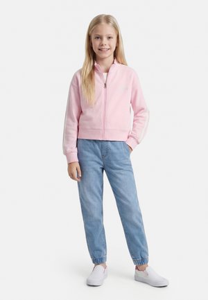 Fată tânără în picioare, purtând o jachetă roz deschis cu fermoar, blugi albaștri cu manșete elastice și pantofi albi slip-on.