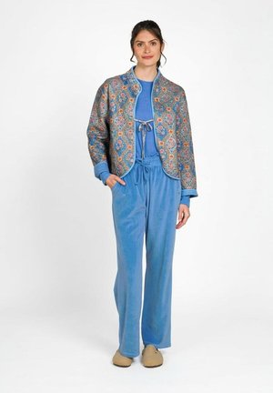 Vrouw staand, gekleed in blauwe veloursbroek en sweater met een kort jasje met bloemenpatroon en beige pantoffels, witte achtergrond.