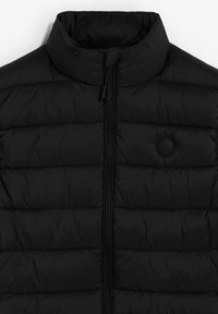 Chaleco acolchado negro con cuello alto, frente con cremallera, textura acolchada y un acento de logo circular. Tejido suave con un acabado mate.