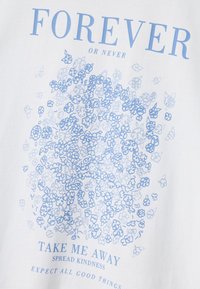 Baltā T-kreklā ir zila grafiskais teksts un ziedu raksts. Teksts ietver "UZMŪŽI VAI NEKAD" un "AIZVEID MANI PROM, IZPLATĪT LABESTĪBU".