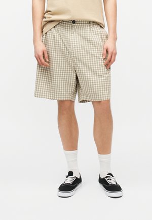 Άνδρας που φοράει μπεζ καρό σορτς, μπεζ πουκάμισο, λευκές κάλτσες και μαύρα αθλητικά παπούτσια, στέκεται μπροστά σε μονόχρωμο φόντο.