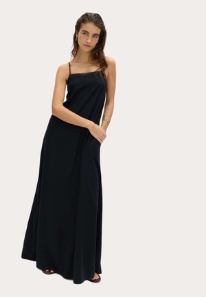 Ung kvinde i en lang sort ærmeløs kjole med tynde stropper står med armene krydset over livet, iført guldbånd og store øreringe.