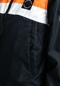 Zwarte jack met een oranje en witte horizontale streep aan de bovenkant. Inclusief een ritssluiting en een knoopzak opzij.