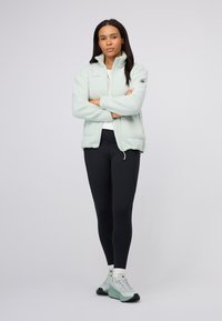 Jasnozielona polarowa kurtka z wysokim kołnierzem i zamek z przodu; zestawiona z czarnymi legginsami i jasnozielonymi butami do wędrówki.