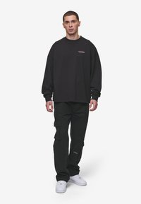 Schwarzer Baumwoll-oversized Sweatshirt mit langen Ärmeln, Rundhalsausschnitt und kleinem Logodetail. Kombiniert mit schwarzen Cargo-Hosen und weißen Sneakern.