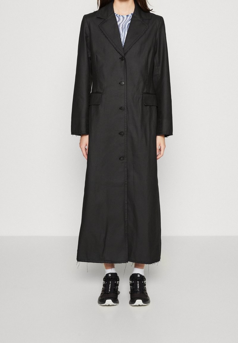 Manteau long noir avec un design structuré, boutonnage à l'avant, col à revers, deux poches latérales et ourlet effiloché. Porté par-dessus un haut rayé et des baskets.