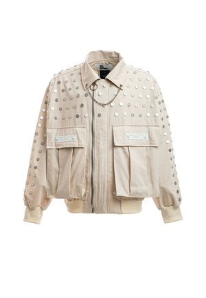 Chaqueta beige con tachuelas circulares plateadas, dos grandes bolsillos delanteros con solapa, cremallera, cuello y una cadena decorativa cruzando el pecho.