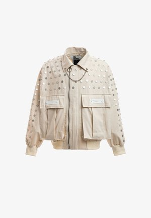 Chaqueta beige con tachuelas circulares plateadas, dos grandes bolsillos delanteros con solapa, cremallera, cuello y una cadena decorativa cruzando el pecho.